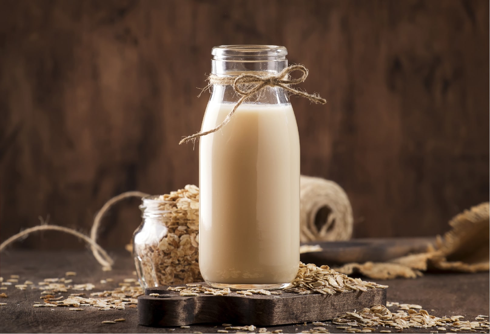 A clear glass bottle of creamy oat milk on a wooden board with scattered oats, evoking natural, gluten-free oat milk