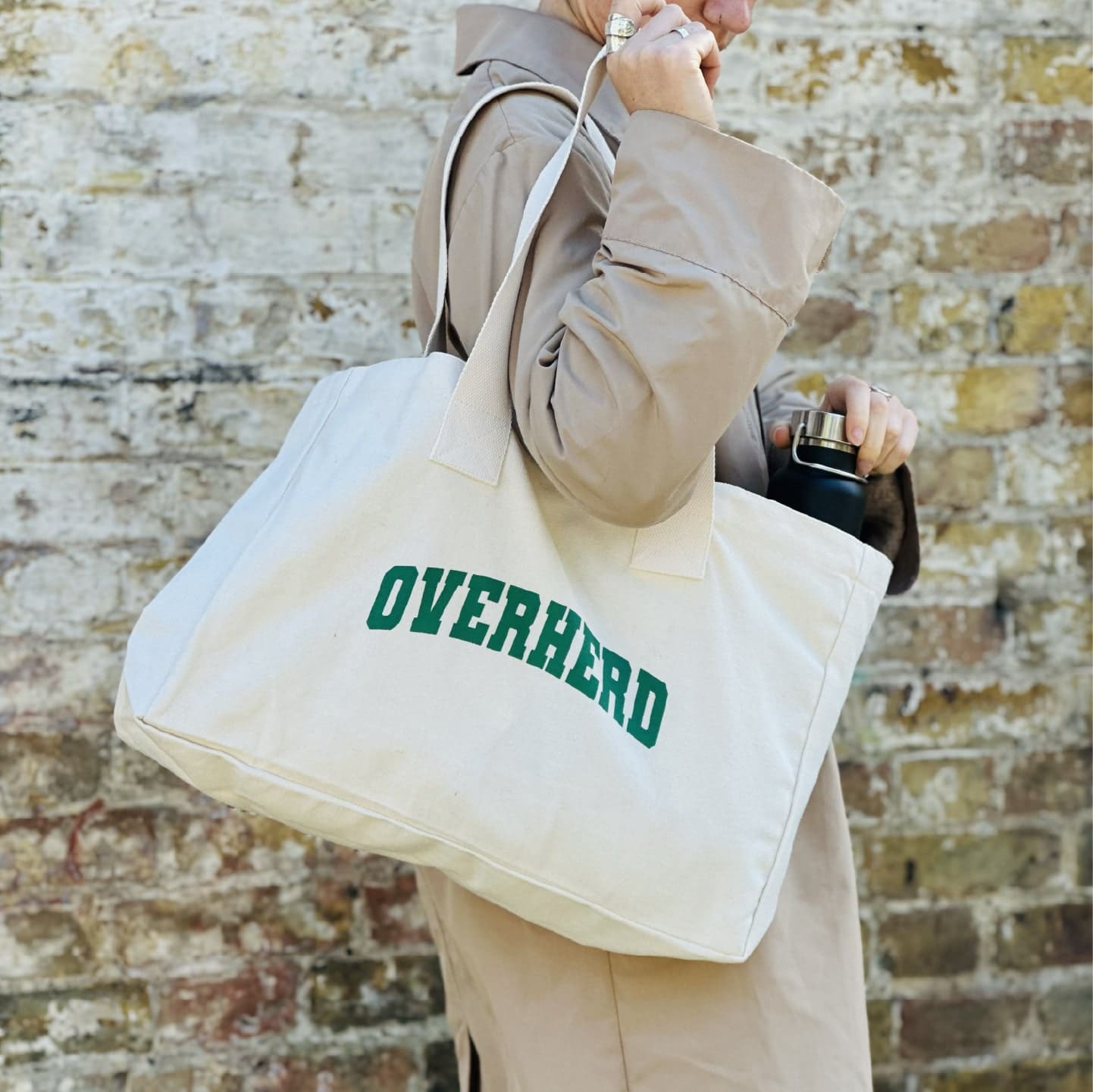 Girl in trench coat holding Overherd tote bag over her shoulder against brick wall background
