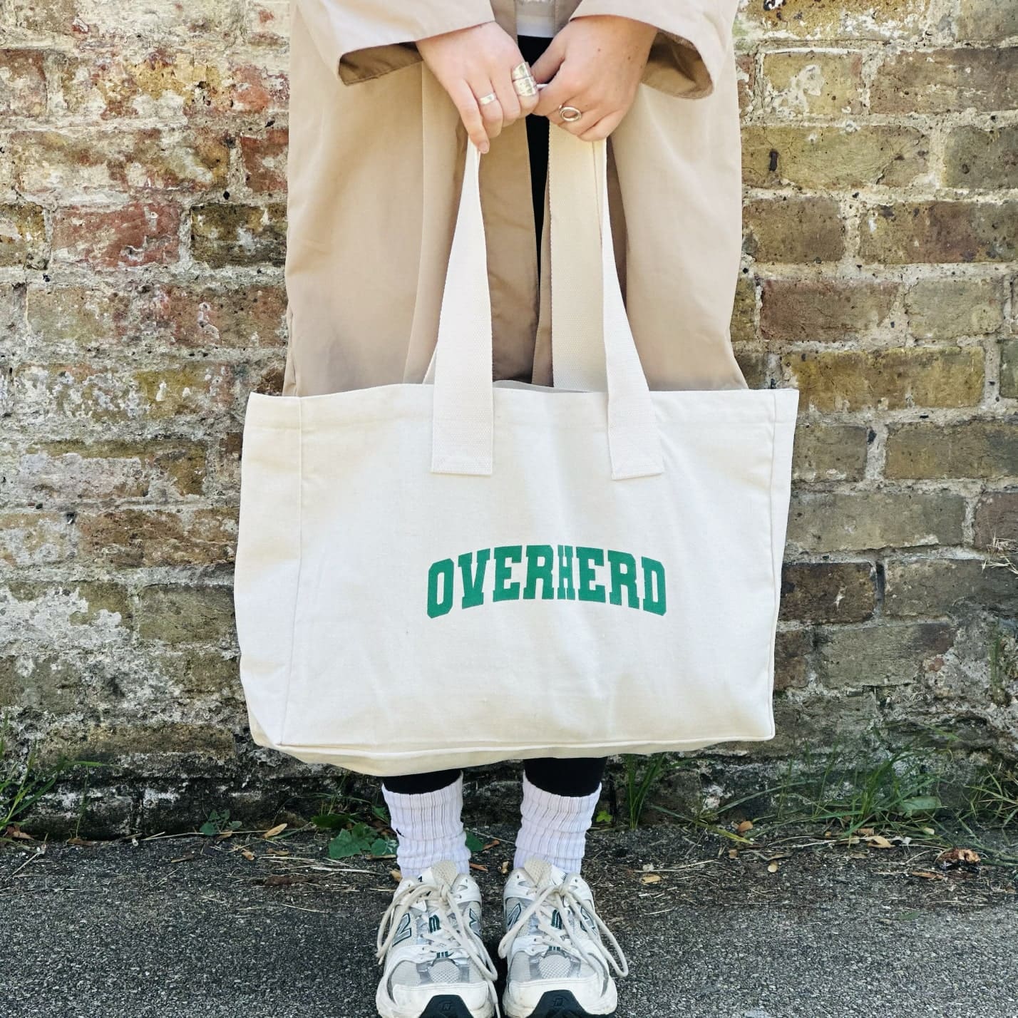 Girl in trench coat holding Overherd tote bag against brick wall background