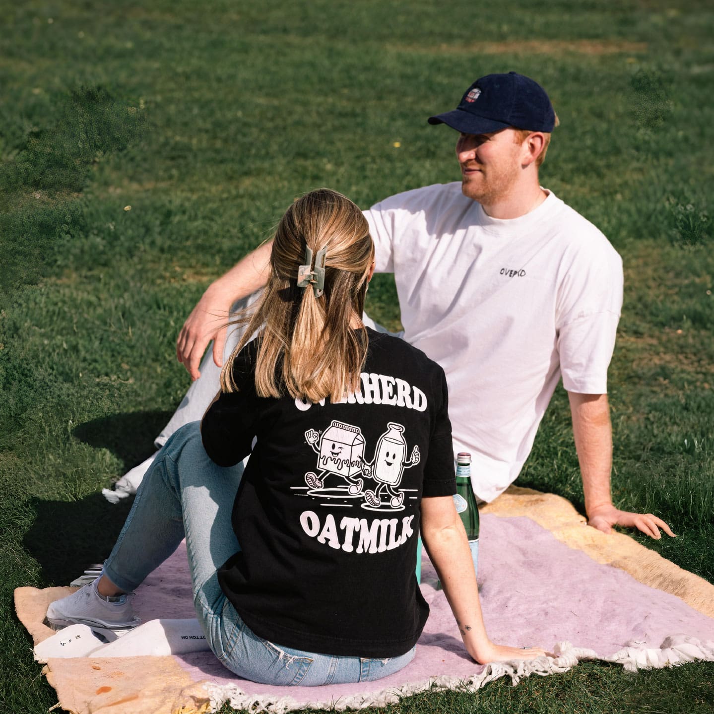 Man and woman sitting on purple blanket wearing oat milk merch