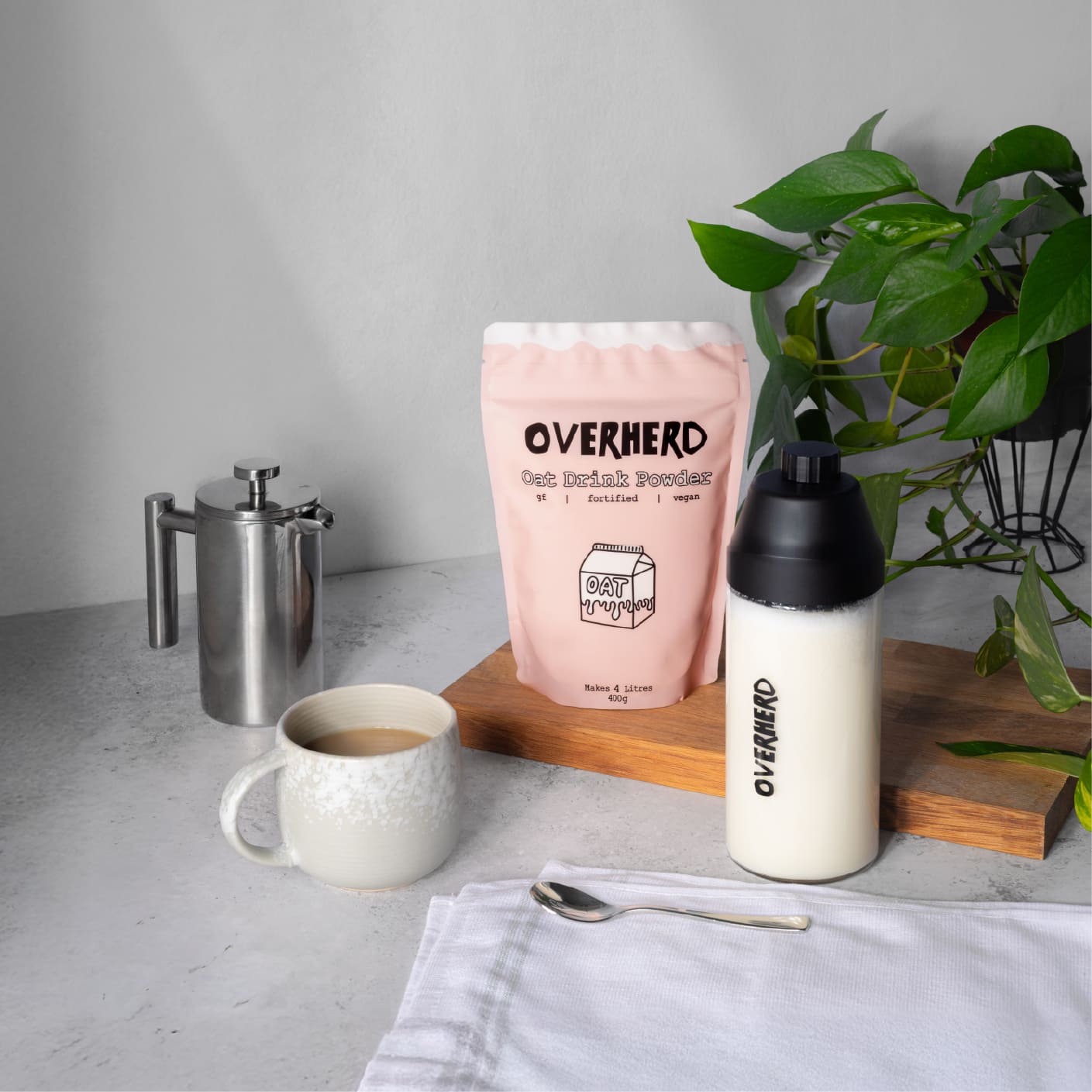Glass bottle of oat milk on table next to mug of coffee and pouch of Overherd oat milk powder