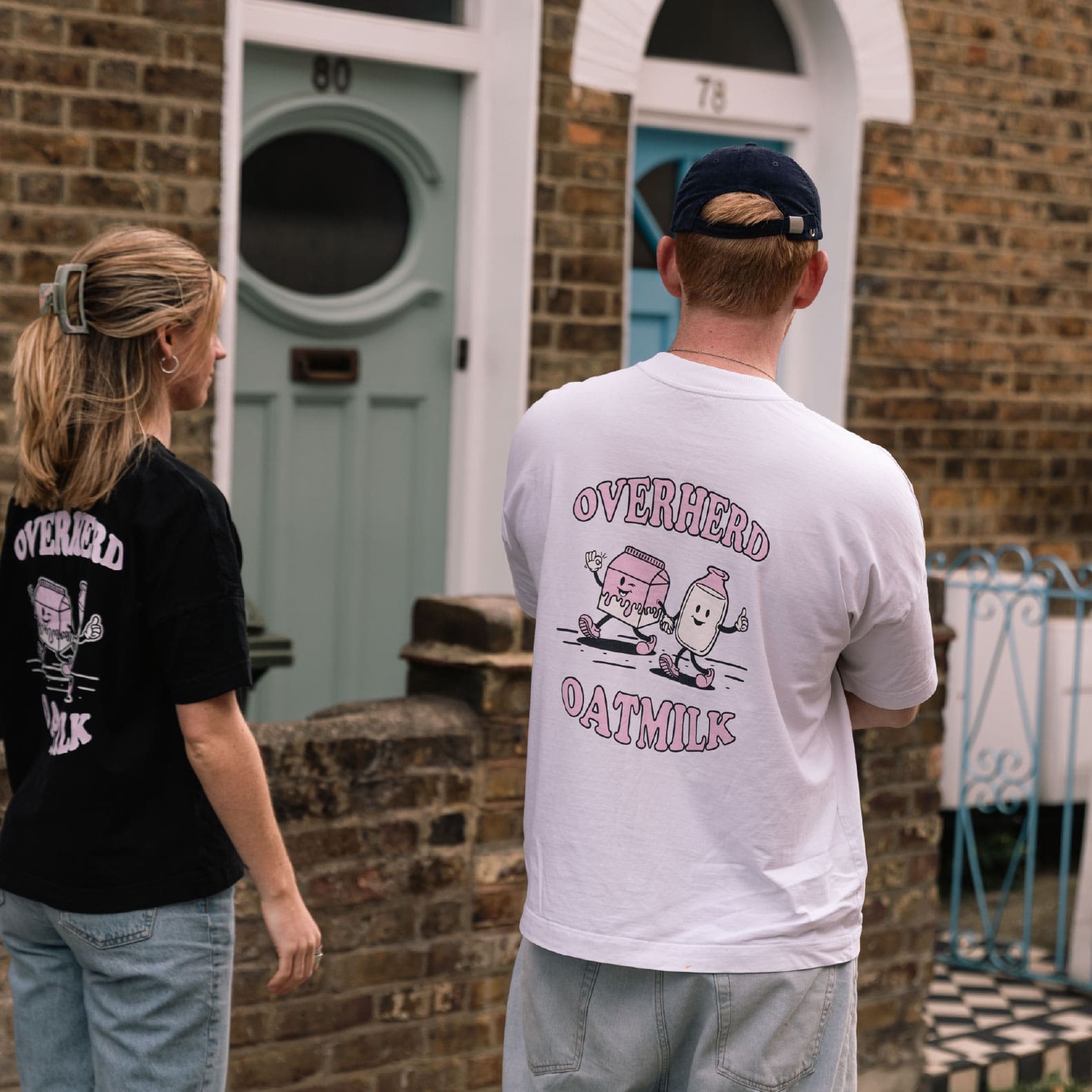 Man and woman wearing oat milk t shirts standing in front of a house